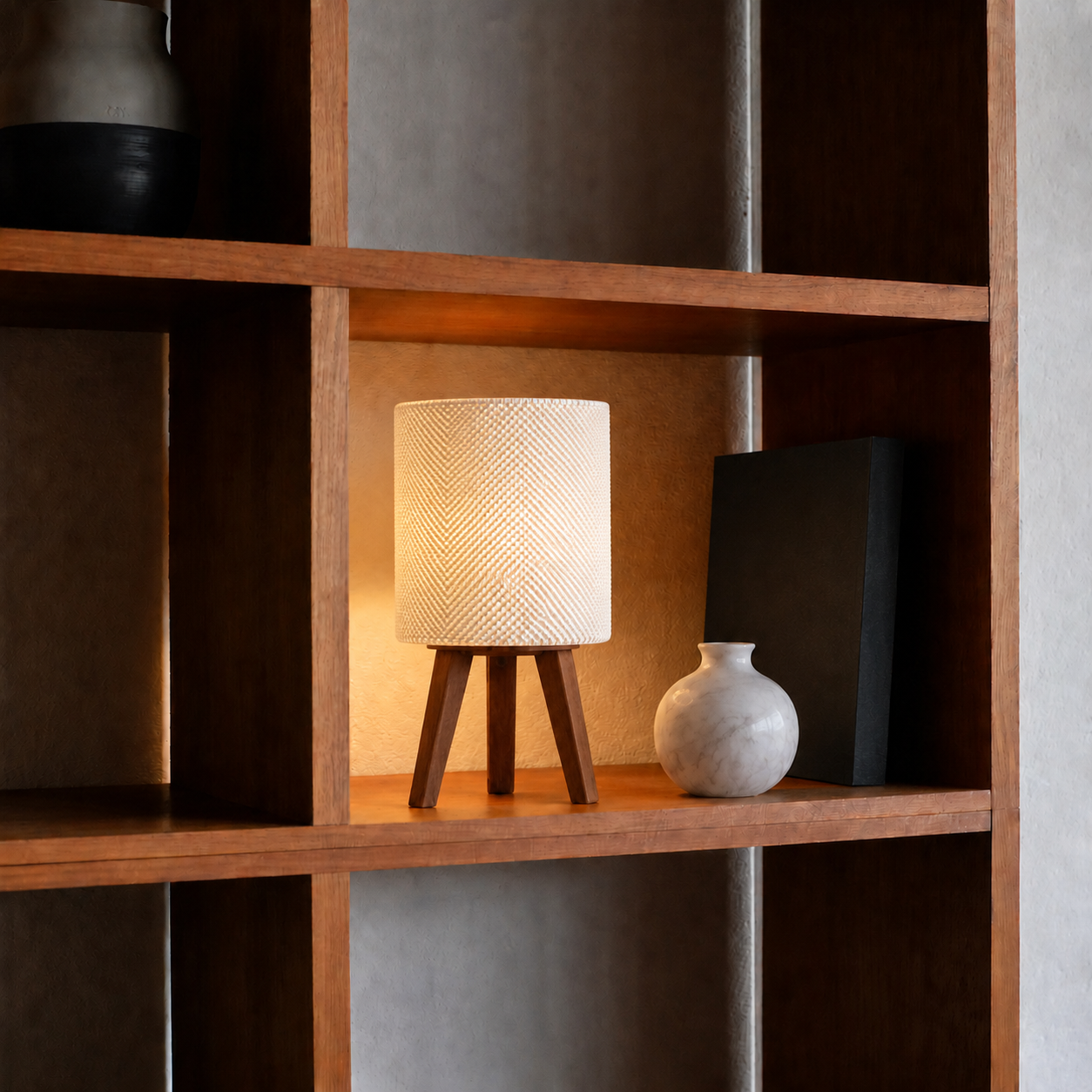 Kete lattice table lamp glowing on a bookshelf next to a white vase, matte white woven shade, LAYR, made in New Zealand